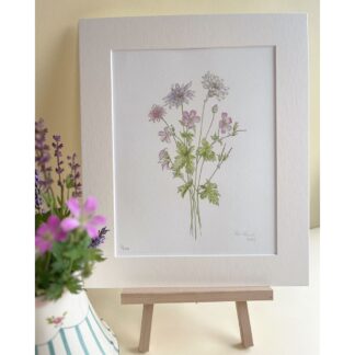 A print of a summer bouquet of flowers in pinks and purples in a mount, standing on an easel. Flowers in a vase in the foreground