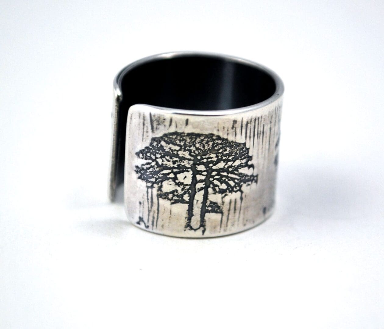 A close-up, slightly angled view of a wide, textured sterling silver cuff ring with an etched tree design on its front, showing the open-back style