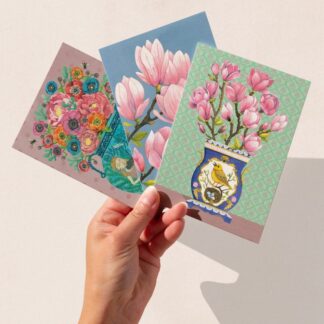 Hand holding a set of three floral art greeting cards with magnolia and vase designs by The Illustrarium, photographed against a soft neutral background.