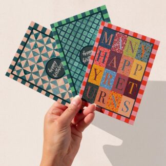 A hand holding three hand-illustrated birthday cards featuring The Illustrarium’s original designs: Many Happy Returns with colourful lettering, and two geometric patterned cards in pink and green with “Happy Birthday” text.