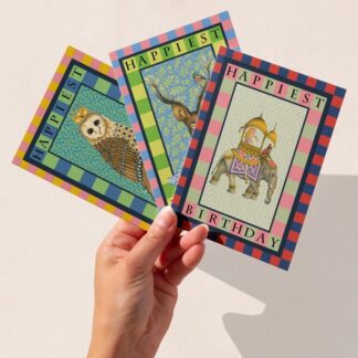 A hand holding three hand-illustrated birthday cards featuring The Illustrarium’s original designs: a crowned owl, a stag with a humorous strong gin banner, and a Indian elephant carrying a girl and a monkey, each bordered in colourful geometric patterns.