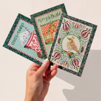 A hand holding three festive Christmas cards featuring The Illustrarium’s original designs: a red mug of hot chocolate reading “Cosy & Warm,” a gingerbread house with “Merry & Bright,” and a crowned robin within a folk-style wreath.