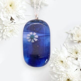 A deep blue glass pendant with a smiling flower, the rear bail reads "Made by Love." It hangs on a silver chain, surrounded by delicate white daisies.