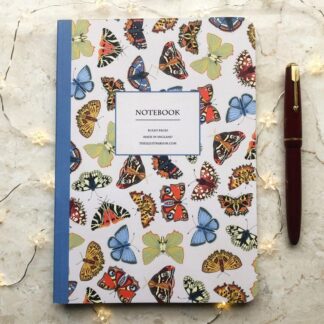 A hand-painted A5 notebook featuring colourful butterflies on a pale beige background, styled with a red fountain pen and soft fairy lights.