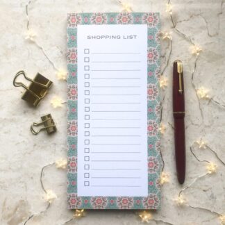 A floral daisy shopping list notepad with a hand-painted green and pink border, gold clips, a red pen, and soft fairy lights on a marble background.