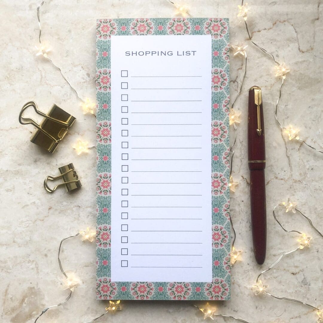 A floral daisy shopping list notepad with a hand-painted green and pink border, gold clips, a red pen, and soft fairy lights on a marble background.