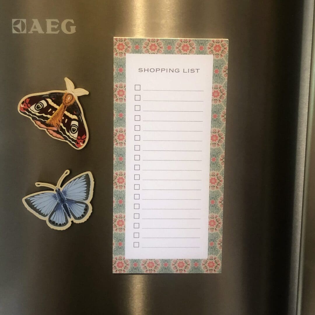 A floral shopping list notepad with a daisy pattern displayed on a stainless steel fridge beside butterfly magnets.