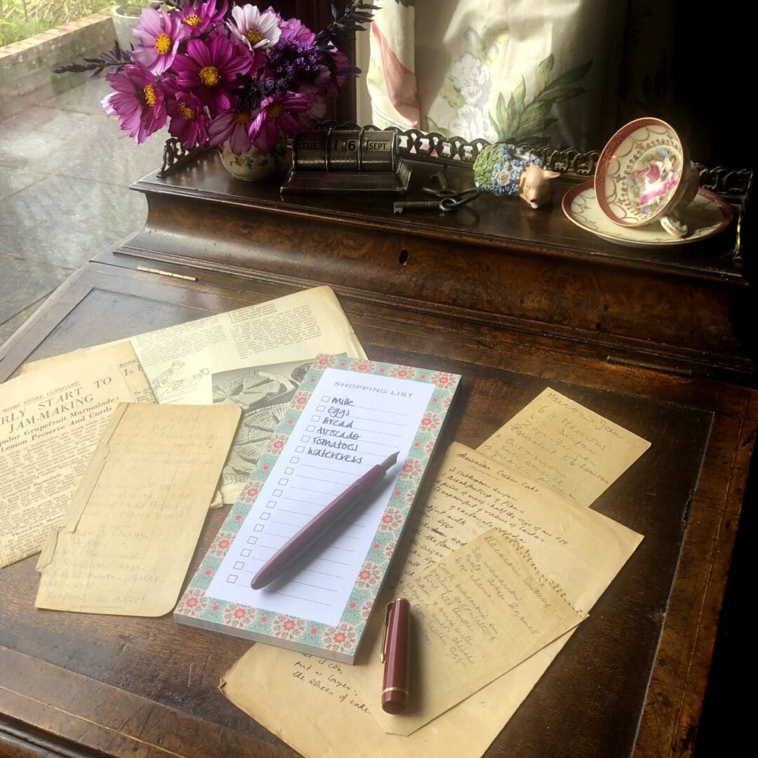 A floral shopping list notepad on a wooden desk surrounded by vintage recipe notes, a red pen, and a vase of purple flowers.