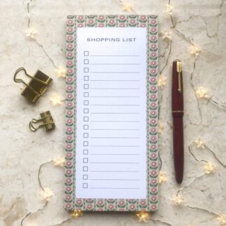 A floral striped shopping list notepad with a pink and green daisy border, gold clips, red pen, and soft fairy lights on a marble background.