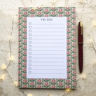 A5 Daisy Stripe notepad featuring hand-painted red and pink daisies on a leafy green patterned background, with a red pen and soft fairy lights beside it.