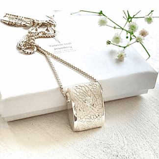 A fine silver pendant, square design with a textured pattern and a white cz stone in the centre. The pendant has a sterling silver chain and is displayed on a white background leaning on a white gift box.
