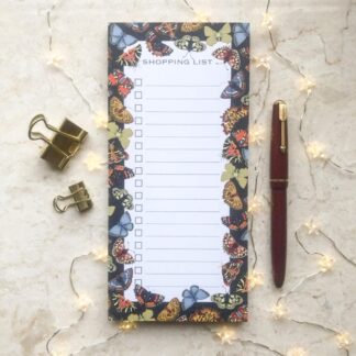 A butterfly shopping list notepad with a dark floral border featuring colourful hand-painted butterflies, styled with gold clips, a red pen, and fairy lights on a marble background.