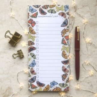 A butterfly shopping list notepad with a bright folk-inspired butterfly border, styled with gold clips, a red pen, and fairy lights on a marble background.
