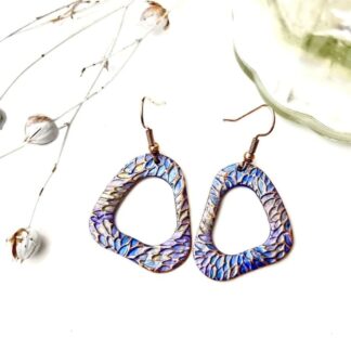 A pair of handcrafted copper drop earrings in a organic hoop shape, they are textured and hand- painted in metallic blues and lilac. Displayed on a white background.