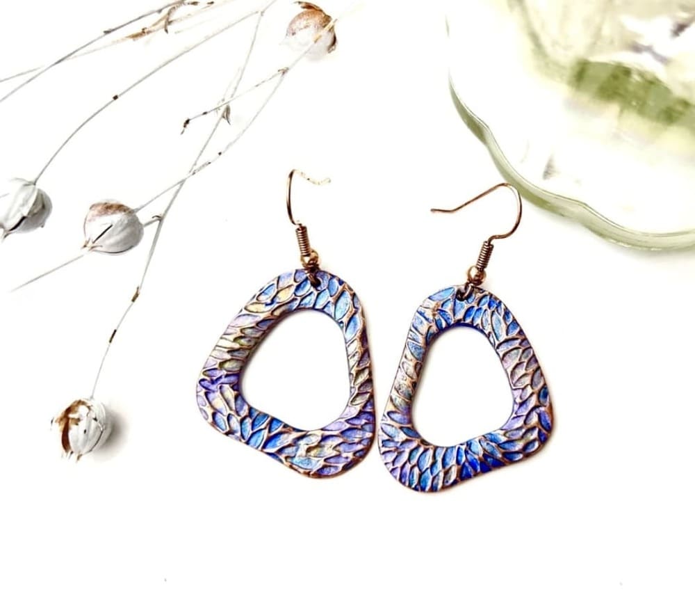 A pair of handcrafted copper drop earrings in a organic hoop shape, they are textured and hand- painted in metallic blues and lilac. Displayed on a white background.