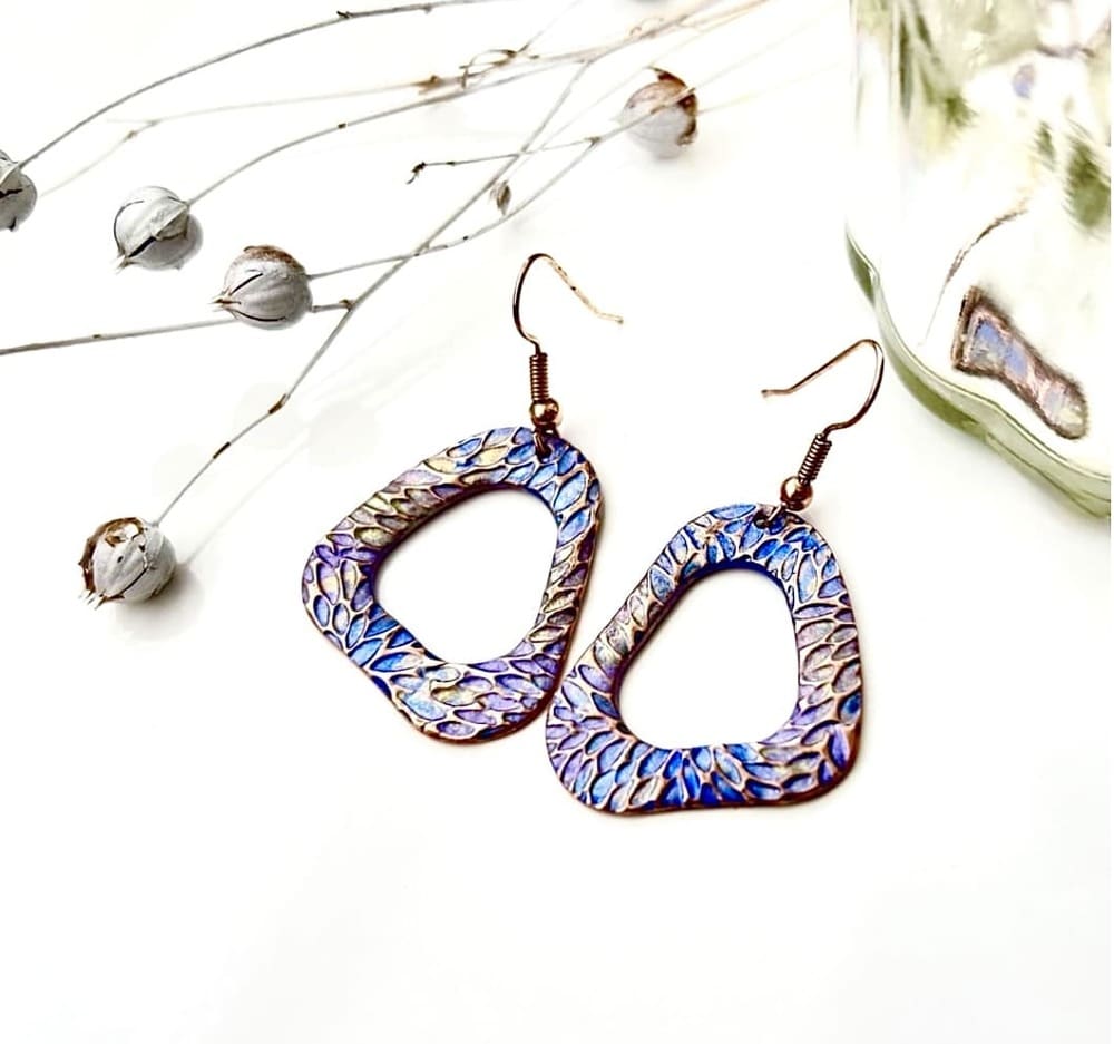 A pair of handcrafted copper drop earrings in a organic hoop shape, they are textured and hand- painted in metallic blues and lilac. Displayed on a white background.