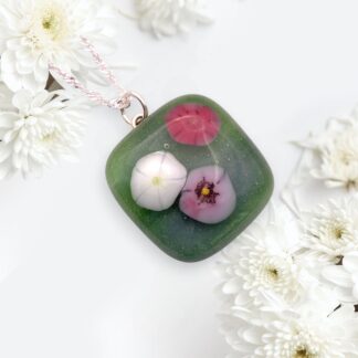 A green handmade glass pendant with delicate pink and white flowers inside, attached to a silver chain, lies among soft white chrysanthemums. Elegant and serene.