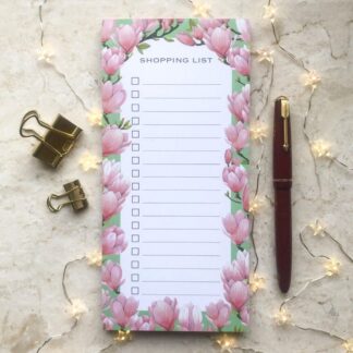 A floral shopping list notepad featuring hand-painted pink magnolia blossoms, styled with gold clips, a red pen, and soft fairy lights on a marble background.