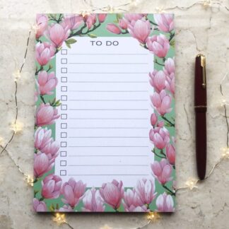 A5 Pink Magnolias notepad featuring hand-painted magnolia flowers on a soft green background, styled with a red pen and delicate fairy lights.