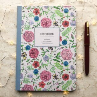 A hand-painted floral A5 notebook with a blue spine and intricate medieval-inspired flower and leaf motifs, styled with fairy lights and a red fountain pen.