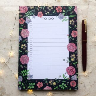 A hand-illustrated A5 notepad with floral designs inspired by medieval manuscripts, featuring a “To Do” layout on recycled paper with a dark background and a fountain pen beside it.