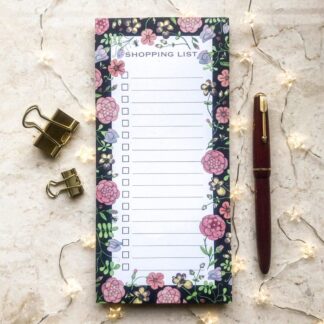 A floral shopping list notepad featuring a dark hand-painted medieval-inspired flower pattern, styled with a red pen, gold clips, and warm fairy lights.