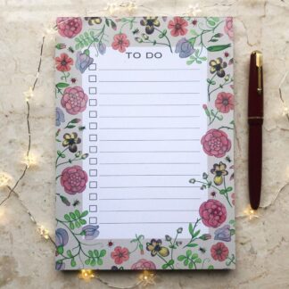 A hand-painted floral A5 to-do notepad with a pale background, featuring pink and red medieval-style flowers, small bees and ladybirds, styled with fairy lights and a fountain pen.