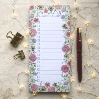 Light floral shopping list notepad with hand-painted medieval-inspired flower design, styled with gold clips, a red pen, and warm fairy lights.