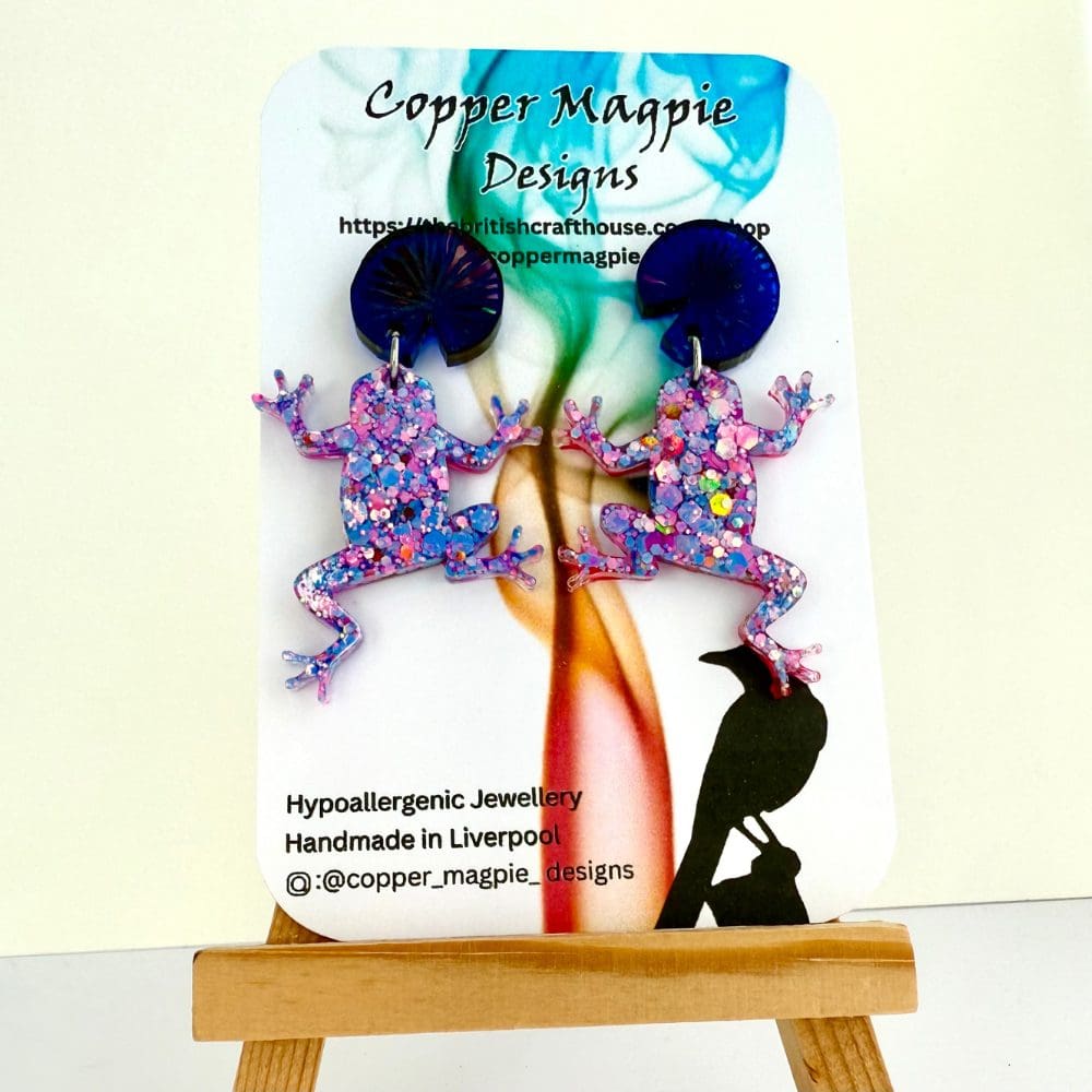 Pink and blue glitter frogs with blue lily pad stud toppers displayed on a branded Copper Magpie Designs card.