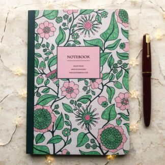 A hand-painted A5 notebook with a pink and green floral pattern inspired by Regency design, styled with a red fountain pen and fairy lights on a marble surface.