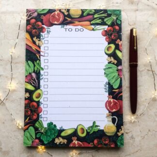 A hand-painted A5 to-do notepad featuring colourful vegetables and fruit — including carrots, avocados, pomegranates, and tomatoes — on a dark background, styled with a red fountain pen and fairy lights.
