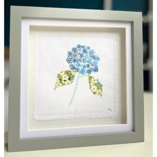 A grey square frame with a single stem blue hydrangea with two green leaves on a white background. Made from applique and embroidery
