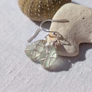 Earrings made of pale green sea-glass wrapped in silver wire, with silver hooks, displayed on a cream coloured cloth and reasting aganst a cream coloured stone with a small piece of sea-urchin showing in the background