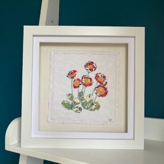 A white square box frame on a shelf featuring a clump of pink daisies appliqued on to white fabric.