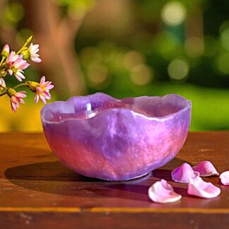An artisanal purple and pink bowl with an irregular edge, sitting on top of a wooden table with pink petals to the right of it and pink flowers to the left