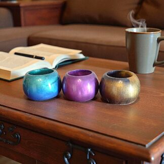 Three round pots, one blue and green, one pink and purple and one black and gold on a wooden table in front of a book and a steaming mug of coffee. In the background the sofa is just visible.