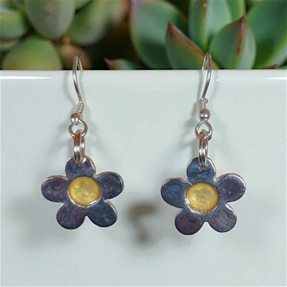 Silver daisy shaped drop earrings handing from a white board in front of succulent plants