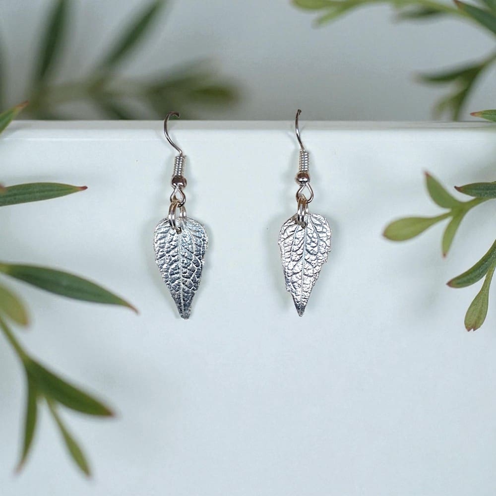 Silver leaf drop earrings hanging from a white block surrounded by leaves