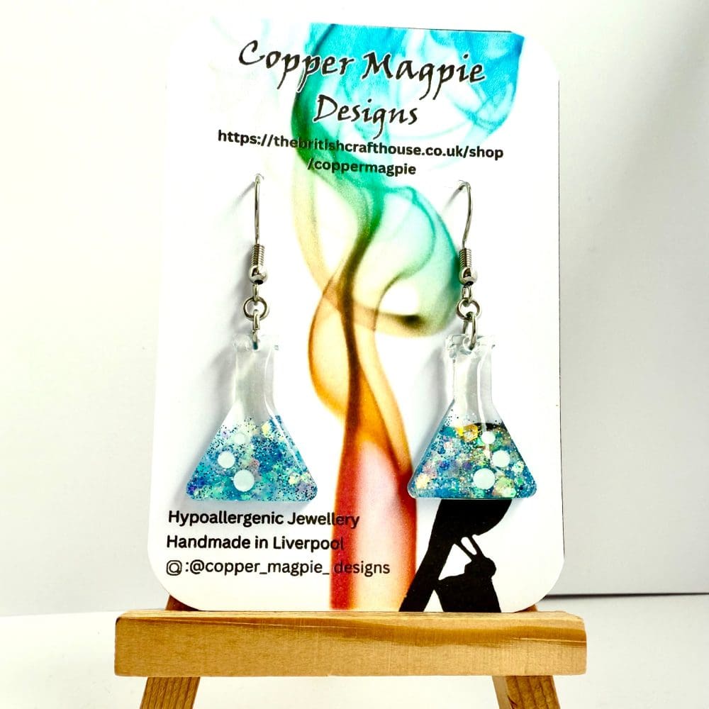 Handmade resin conical flask earrings in blue glitter named Neptune’s Tonic hanging on hypoallergenic surgical steel hooks, displayed on a branded Copper Magpie Designs earring card.
