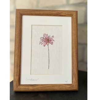 A wooden frame standing in front of a white wall featuring a single pink embroidered cornflower.