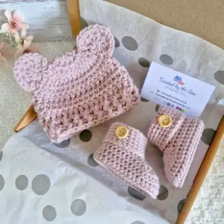 A handmade crochet baby hat and matching booties set, each featuring a round wooden button engraved with 'Born in 2026' and displayed in a gift box.