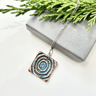 A square shaped copperpendant that has a circle swirl pattern in the centre hand-painted in blues and green. It has a sterling silver fine chain and is displayed on a white background with a grey jewellery box and some fern.