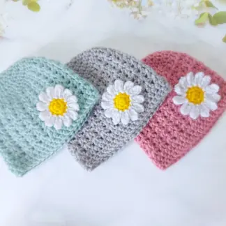 A soft duck egg, silver and pale rose crochet baby beanie hat featuring a small yellow and white daisy motif on the front.