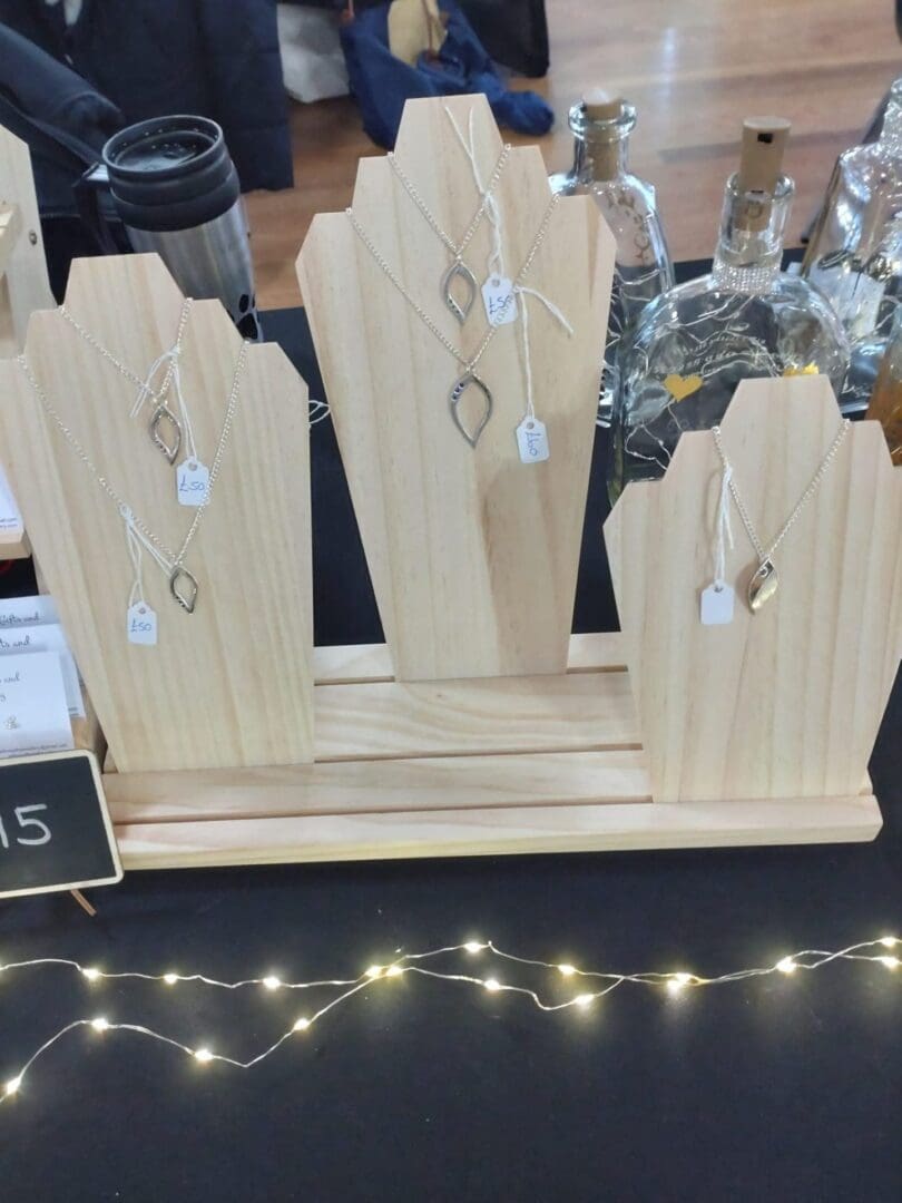 Three wooden jewellery stands on a market stall on top of a black table cloth. Each stand has two or three silver necklaces on them and there are fairy lights in front of the stands