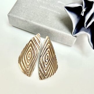 A pair of large brinze stud earrings in a geometric shape. They are displayed on a white background with a grey gift box above.