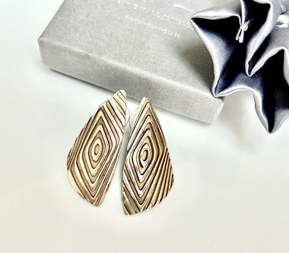 A pair of large brinze stud earrings in a geometric shape. They are displayed on a white background with a grey gift box above.