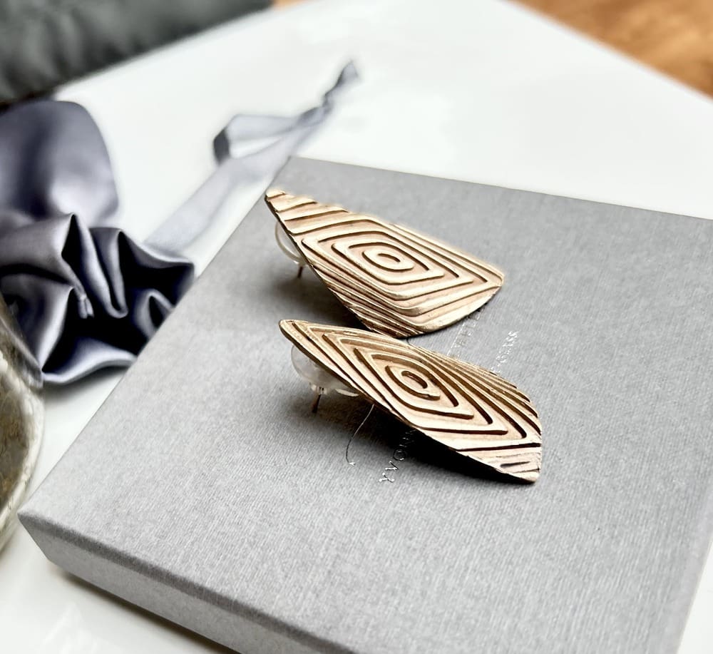 A pair of large brinze stud earrings in a geometric shape. They are displayed sat on a grey jewellery presentation box, looking at them from the side.