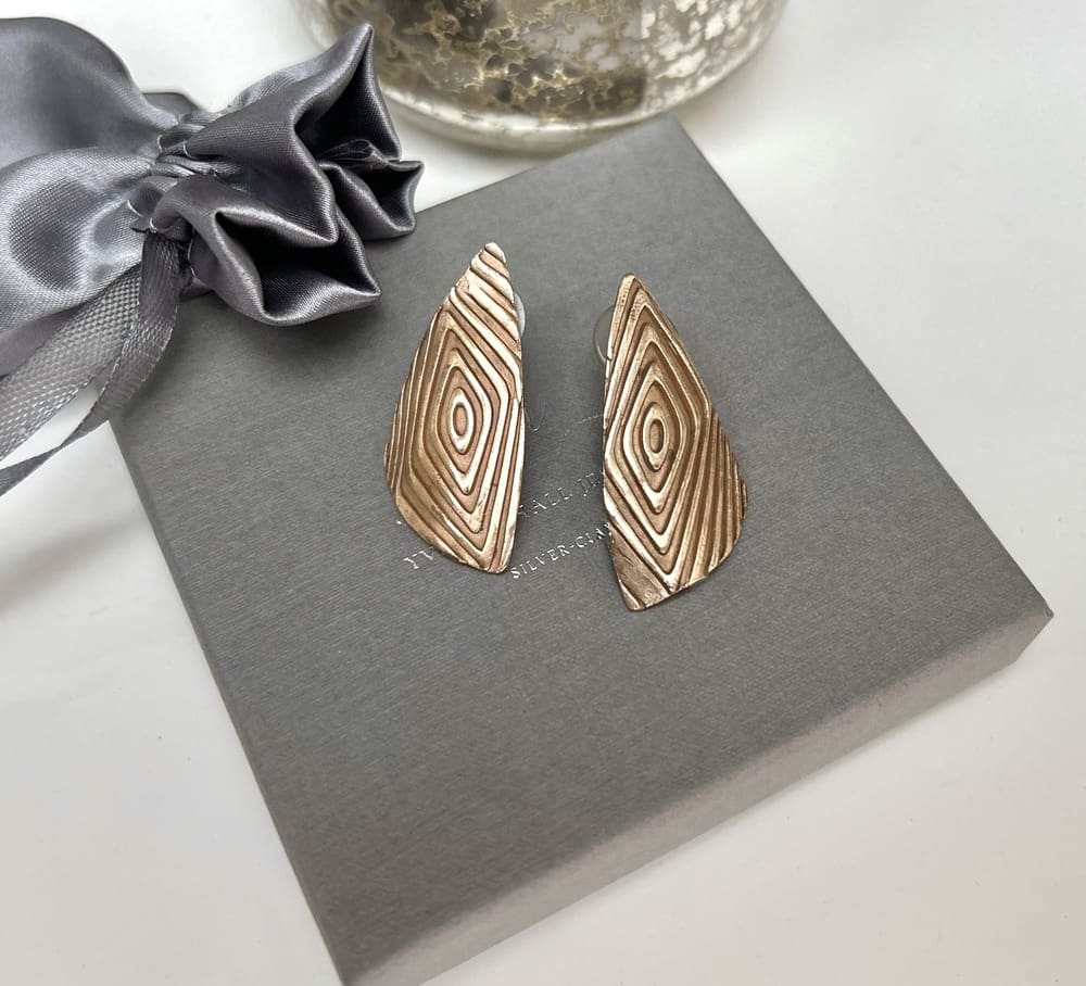 A pair of large brinze stud earrings in a geometric shape. They are displayed sat on a grey jewellery presentation box.