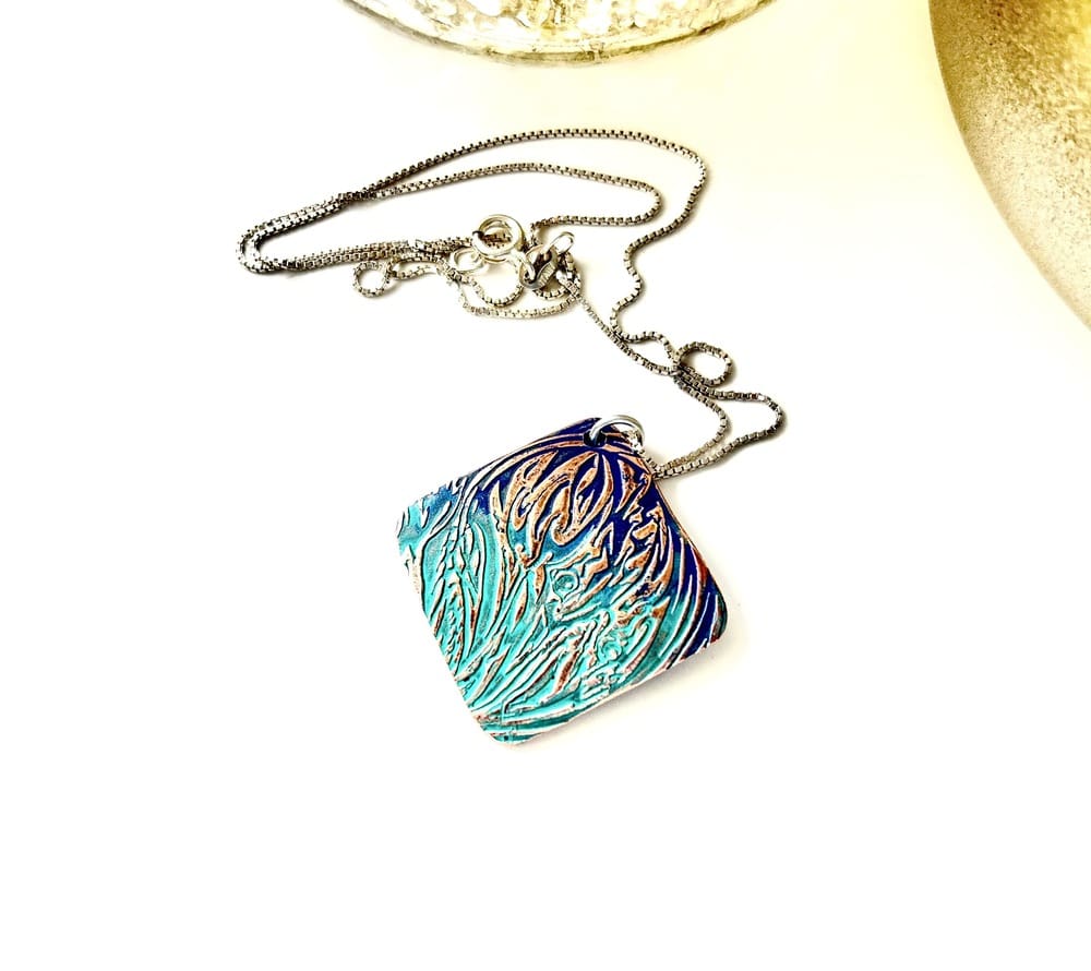 A handcrafted textured copper pendant which has a blue and green patina, it is in a diamond shape and has a sterling silver chain. The pendant is displayed on a white background.