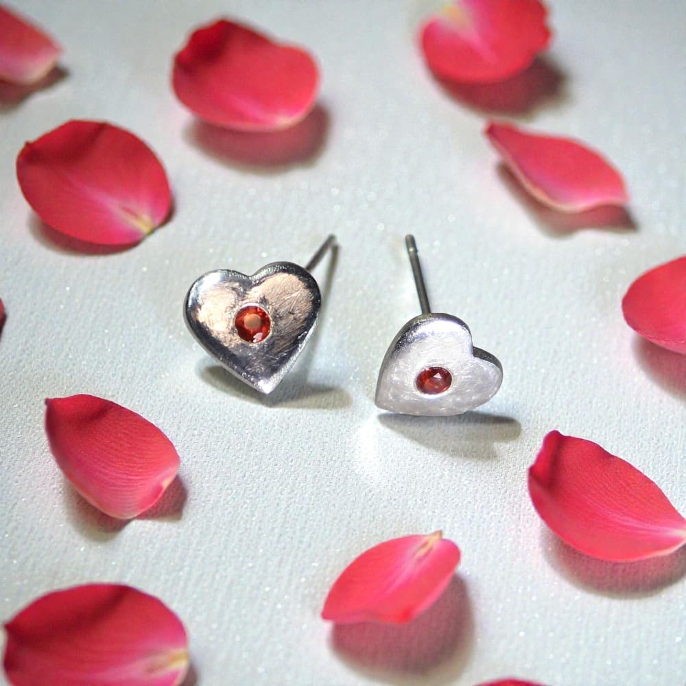 A pair of silver heart shaped stud earring with red stones in the middle of each. Sitting on a white surface, the left one is resting on its point and the right one is resting on its side. Surrounding the earrings are lots of red rose petals